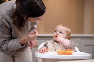 Woman looking at toddler who is eating a tray of food in a chair looking happy