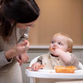 woman looking at toddler who is eating a tray of food in a chair looking happy.