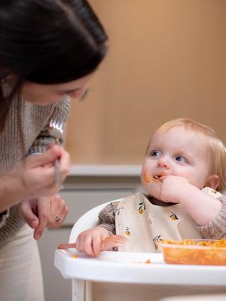 woman looking at toddler who is eating a tray of food in a chair looking happy.