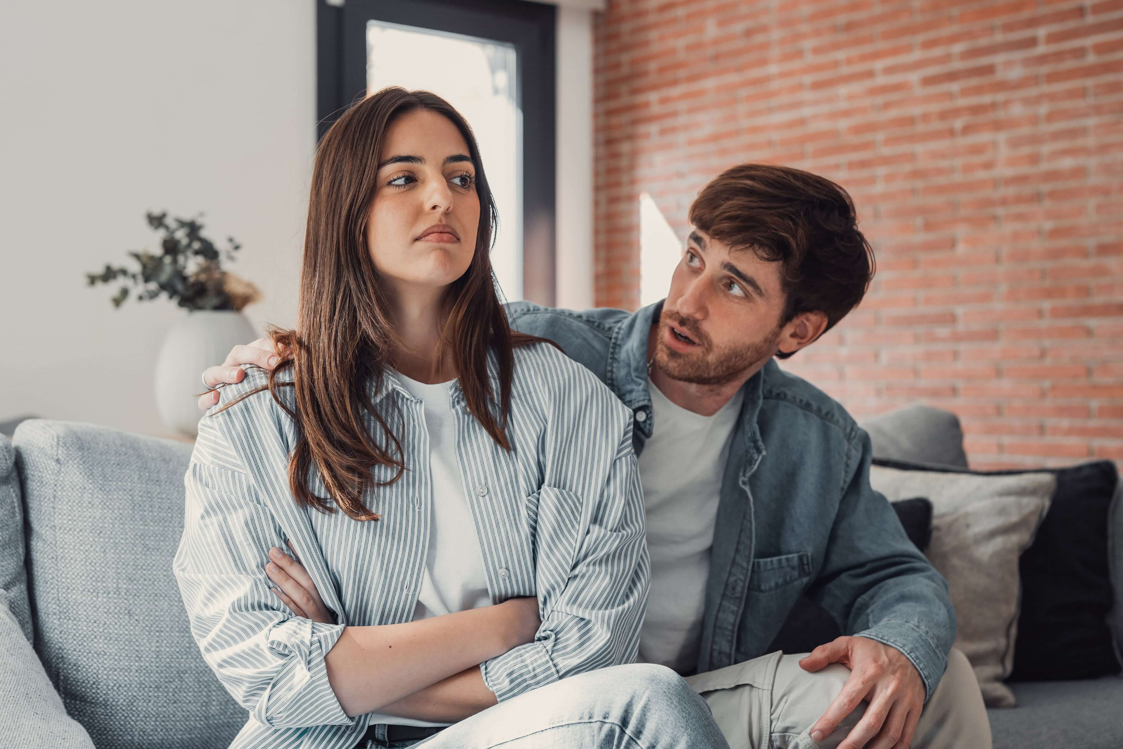 Ein Mann und eine Frau in einem Wohnzimmer, wobei der Mann versucht, mit der Frau zu kommunizieren, die einen strengen Gesichtsausdruck hat.