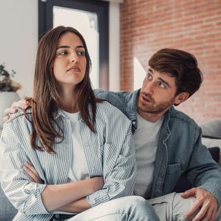 Ein Mann und eine Frau in einem Wohnzimmer, wobei der Mann versucht, mit der Frau zu kommunizieren, die einen strengen Gesichtsausdruck hat.