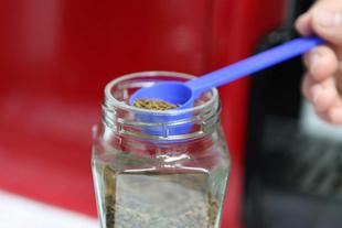 Blauer Milchpulverlöffel portioniert Kaffeepulver aus einem Glasbehälter vor rotem Küchenhintergrund.