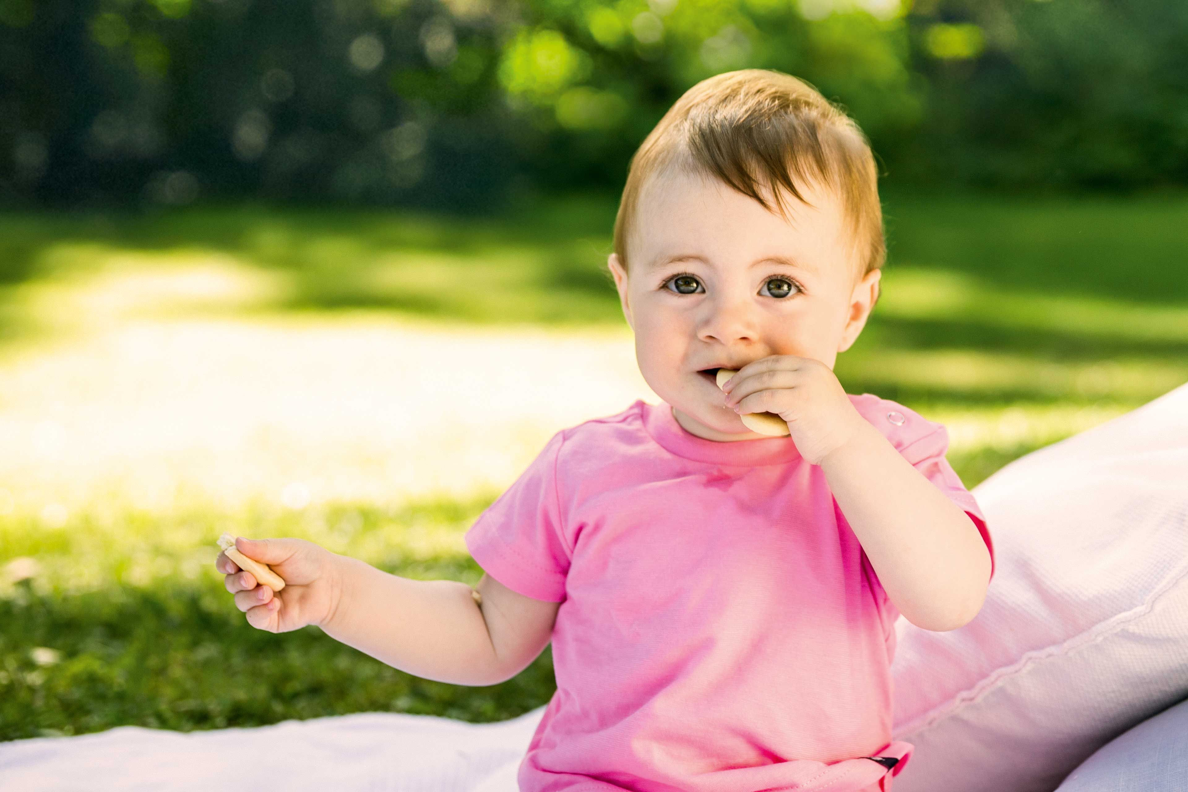 Ein kleines Kind, vermutlich ein Baby, trägt ein rosa Hemd und kaut auf einem grünen Gegenstand, vor einem Hintergrund aus grünem Gras und einem weißen Kissen.