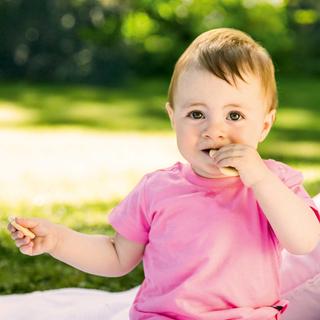 Ein kleines Kind, vermutlich ein Baby, trägt ein rosa Hemd und kaut auf einem grünen Gegenstand, vor einem Hintergrund aus grünem Gras und einem weißen Kissen.
