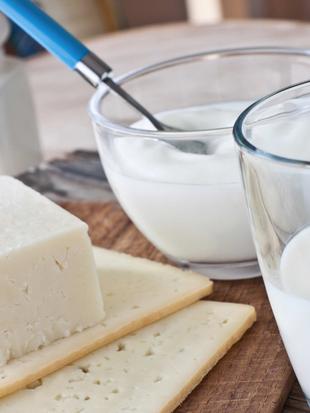 Verschiedene Molkereiprodukte, darunter ein Stück Käse, ein Glas Milch und ein Behälter mit Joghurt, die alle auf einer Holzunterlage platziert sind.
