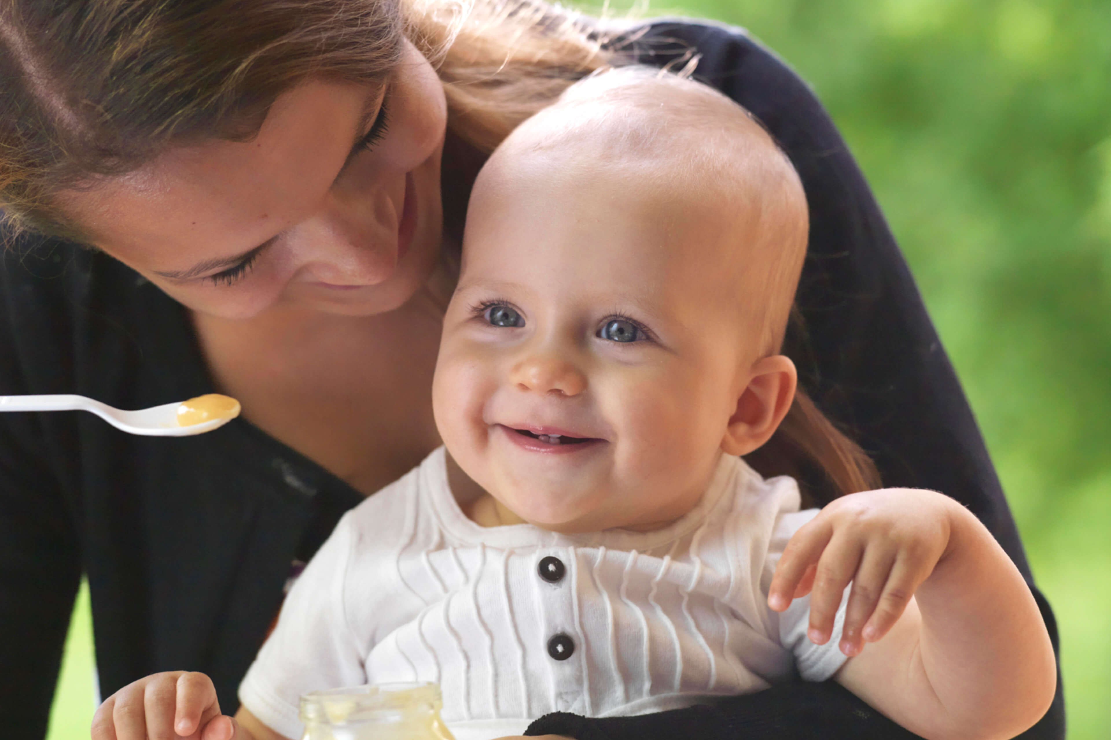 Ein fröhliches Baby mit blauen Augen, das lächelt und in die Kamera schaut, während es von einer Frau gehalten wird, die das Baby mit einem Löffel füttert.