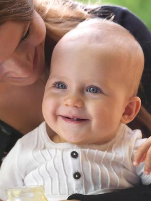 Ein fröhliches Baby mit blauen Augen, das lächelt und in die Kamera schaut, während es von einer Frau gehalten wird, die das Baby mit einem Löffel füttert.