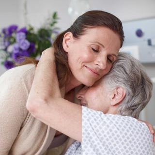 Jüngere Frau umarmt liebevoll eine ältere Patientin im Krankenhausbett