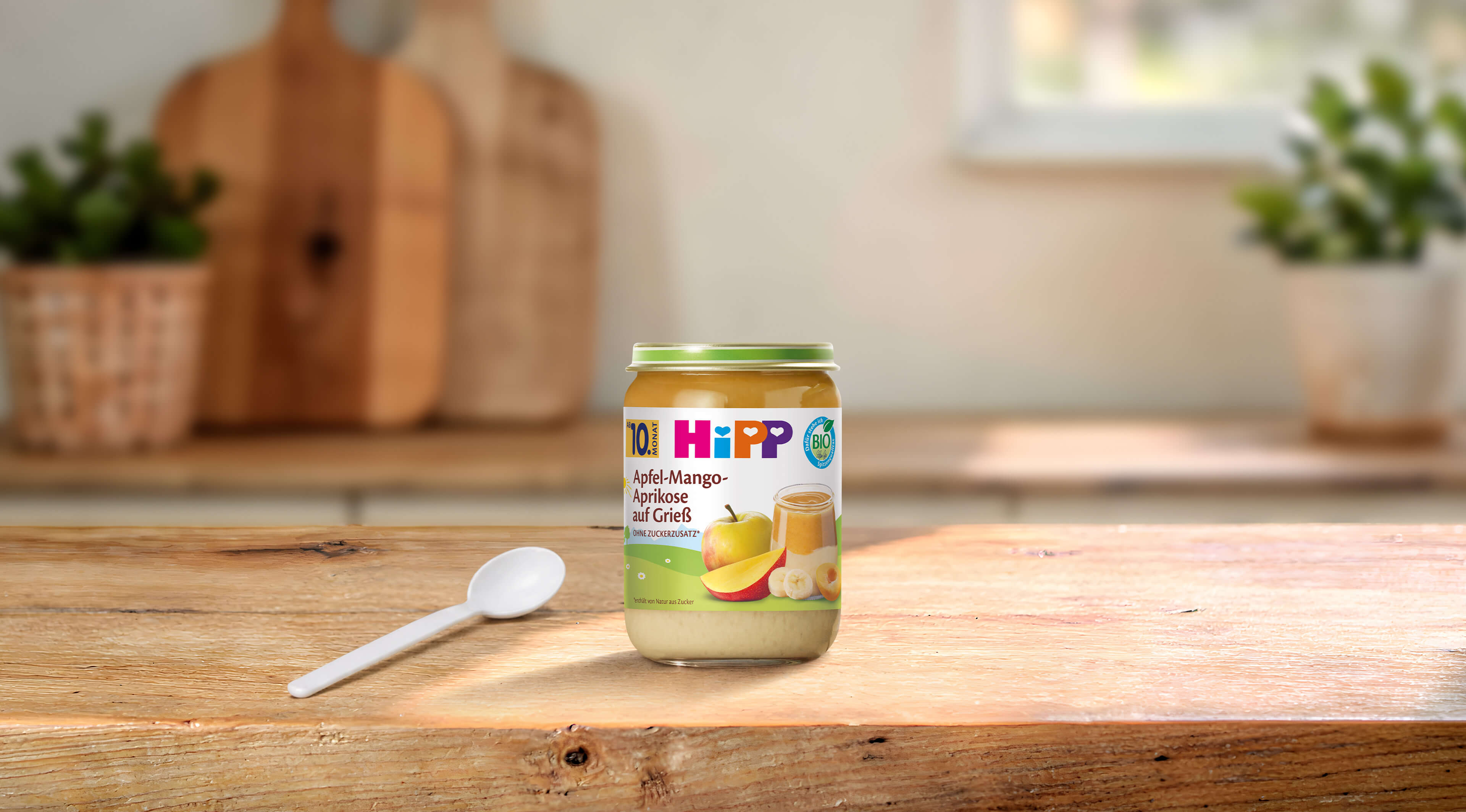 HiPP Bio-Gläschen Apfel-Mango-Aprikose auf Grieß ab 10. Monat auf Holztisch mit Löffel in heller Küche