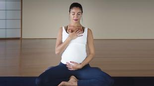 Eine Frau in meditativer Haltung, die ein weißes Tanktop und blaue Leggings trägt, mit geschlossenen Augen und Händen auf Brust und Bauch.