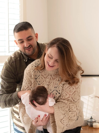 A joyful couple interacts with their baby in a sunlit room. The mother, wearing a cozy knit sweater, holds the baby in her arms while laughing, as the father, dressed in a plaid shirt, leans in with a warm smile. Sunlight streams through white shutters, casting a soft glow on the scene, with a framed bird illustration and decorative items in the background.