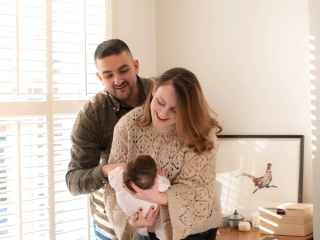 A joyful couple interacts with their baby in a sunlit room. The mother, wearing a cozy knit sweater, holds the baby in her arms while laughing, as the father, dressed in a plaid shirt, leans in with a warm smile. Sunlight streams through white shutters, casting a soft glow on the scene, with a framed bird illustration and decorative items in the background.