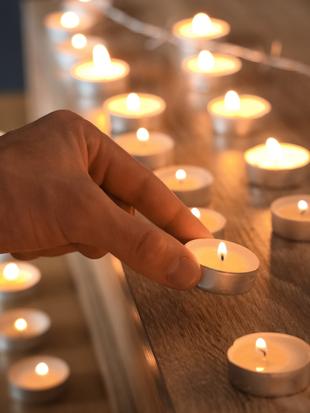 Eine Hand, die eine brennende Teelichtkerze auf eine hölzerne Oberfläche stellt, umgeben von anderen Kerzen.