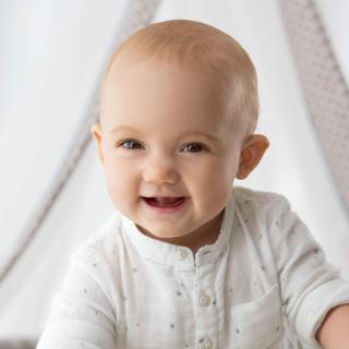 Ein Baby mit einem freudigen Gesichtsausdruck, das ein weißes Hemd trägt, vor einem weichen Hintergrund.
