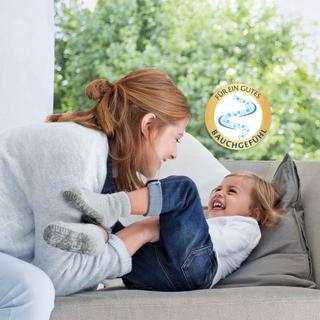 Lachende Mutter kitzelt ihr Kleinkind auf dem Sofa, daneben ein Siegel mit dem Text „Für ein gutes Bauchgefühl“ vor grünem Hintergrund.