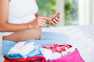 Eine schwangere Frau sitzt auf einem Bett, hält ein Mobiltelefon in der Hand und hat eine rosa Tasche mit Babyartikeln dabei.