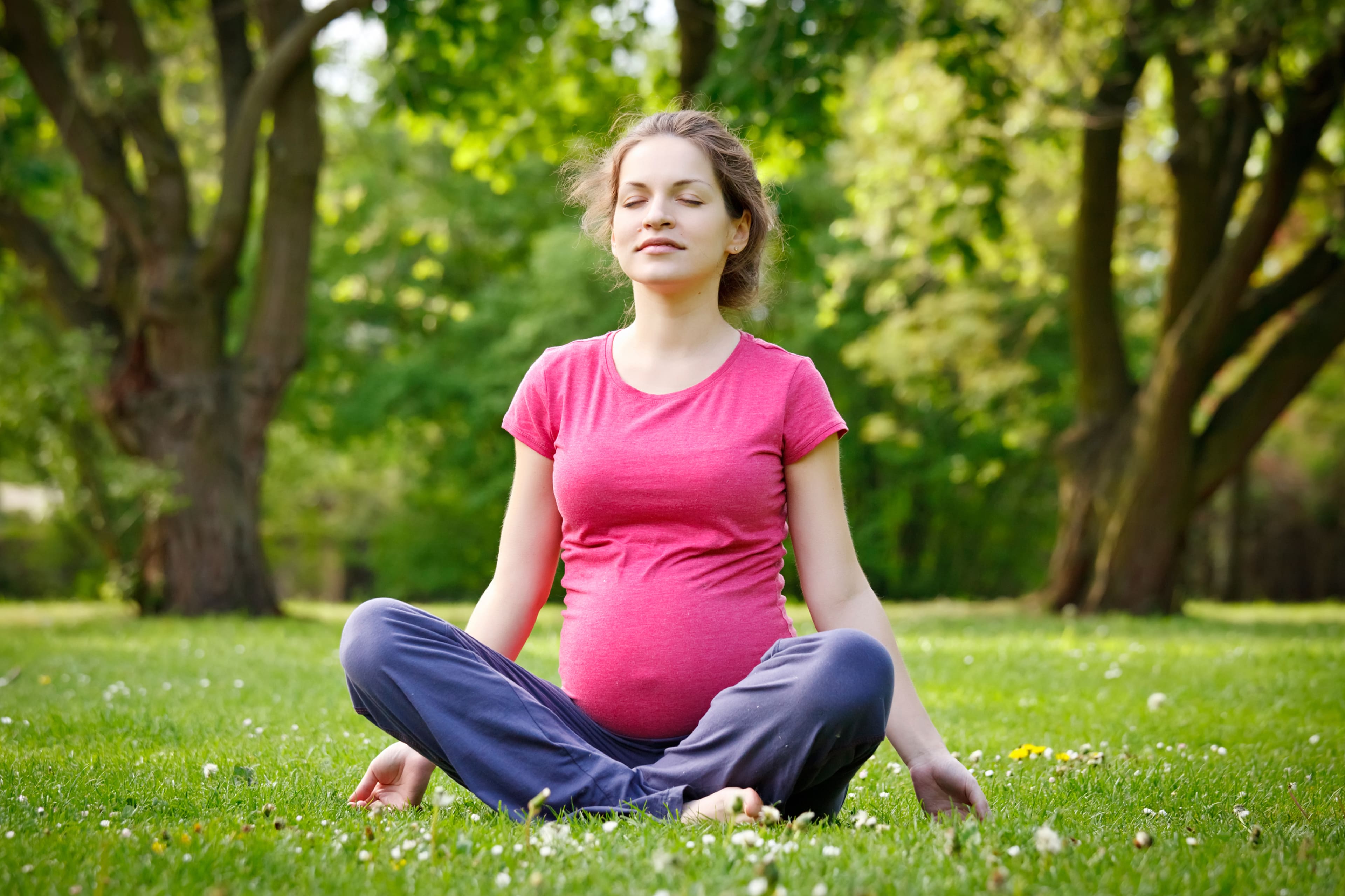 Eine Frau in einem rosa Hemd und einer blauen Hose sitzt in meditativer Haltung auf einer von Bäumen umgebenen Wiese.