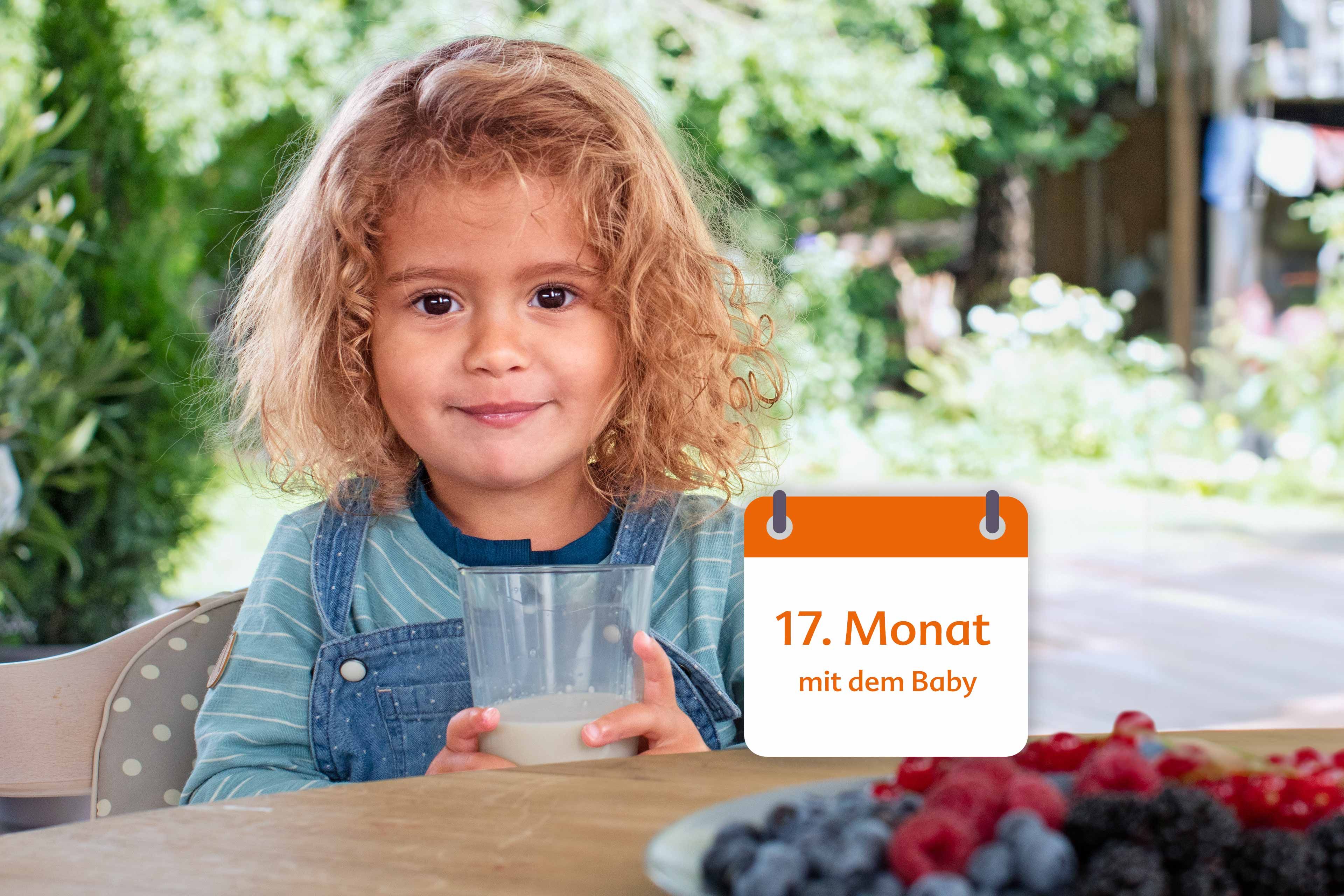 Ein kleines Kind mit lockigem Haar, das ein Glas Milch in der Hand hält, vor einem Gartenhintergrund mit einem Kalender, auf dem "17. Monat mit dem Baby".