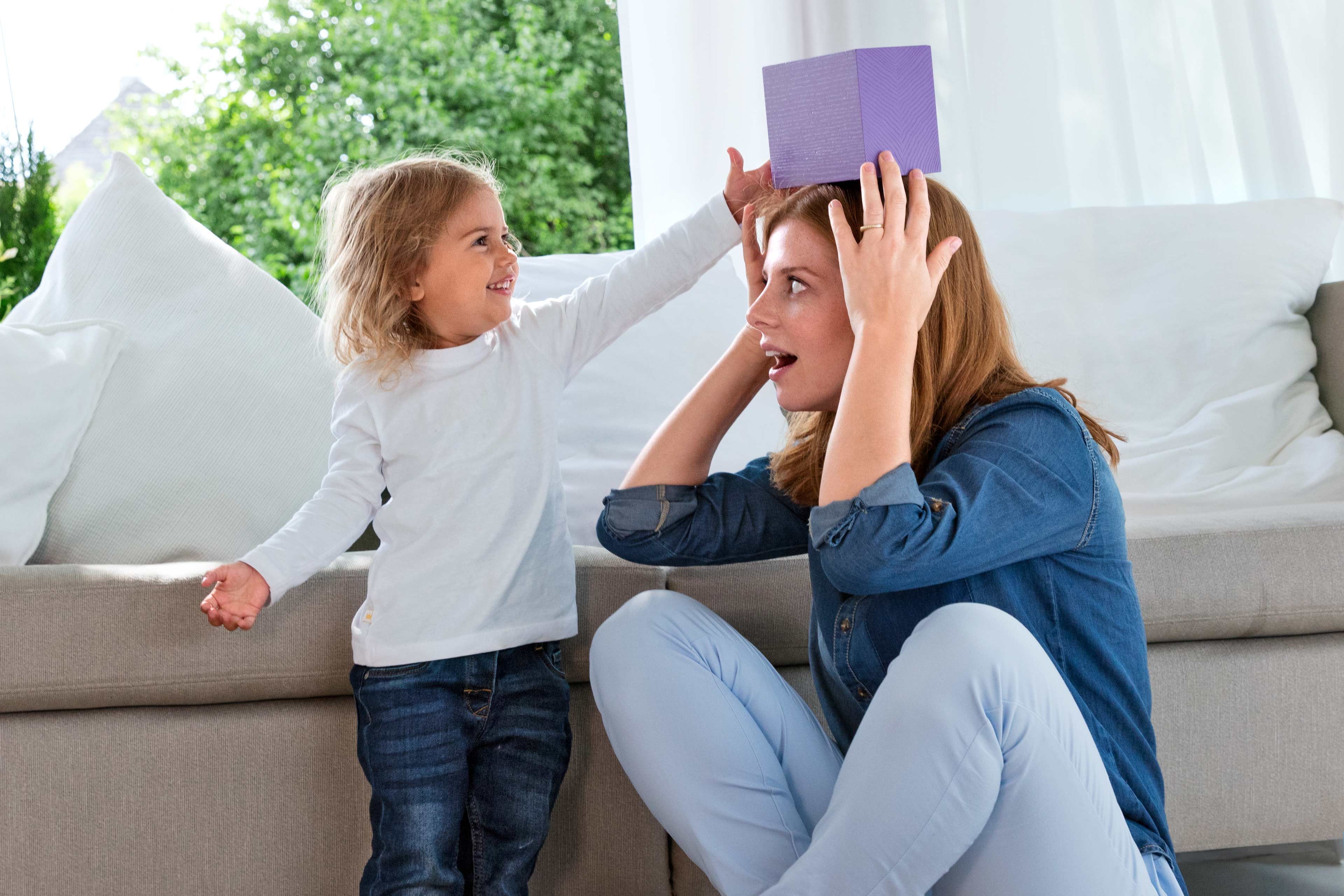 Ein junges Mädchen setzt einer Frau spielerisch eine lila Schachtel auf den Kopf, während die Frau überrascht aussieht.