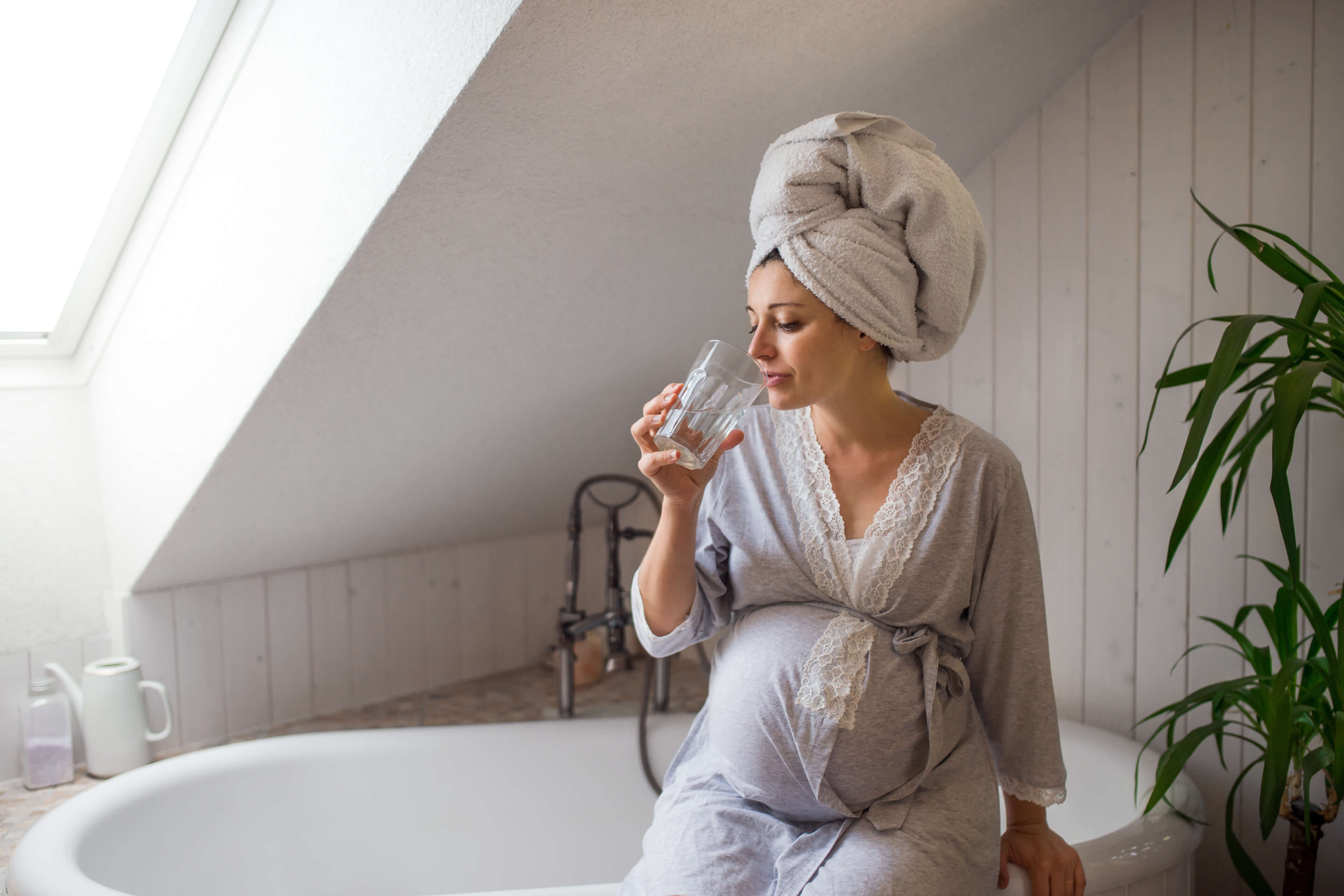 Eine Frau in einem Badezimmer, in Bademantel und Handtuch, die Wasser aus einem Glas trinkt.