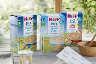 Drei Schachteln mit Babynahrung mit den Aufschriften "BIO MILCHBREI", "BIO MILCHBREI Kinder-Gries" und "BIO MILCHBREI Hafer Apfel", jede mit einer anderen Geschmacksrichtung und Nährwertangaben.