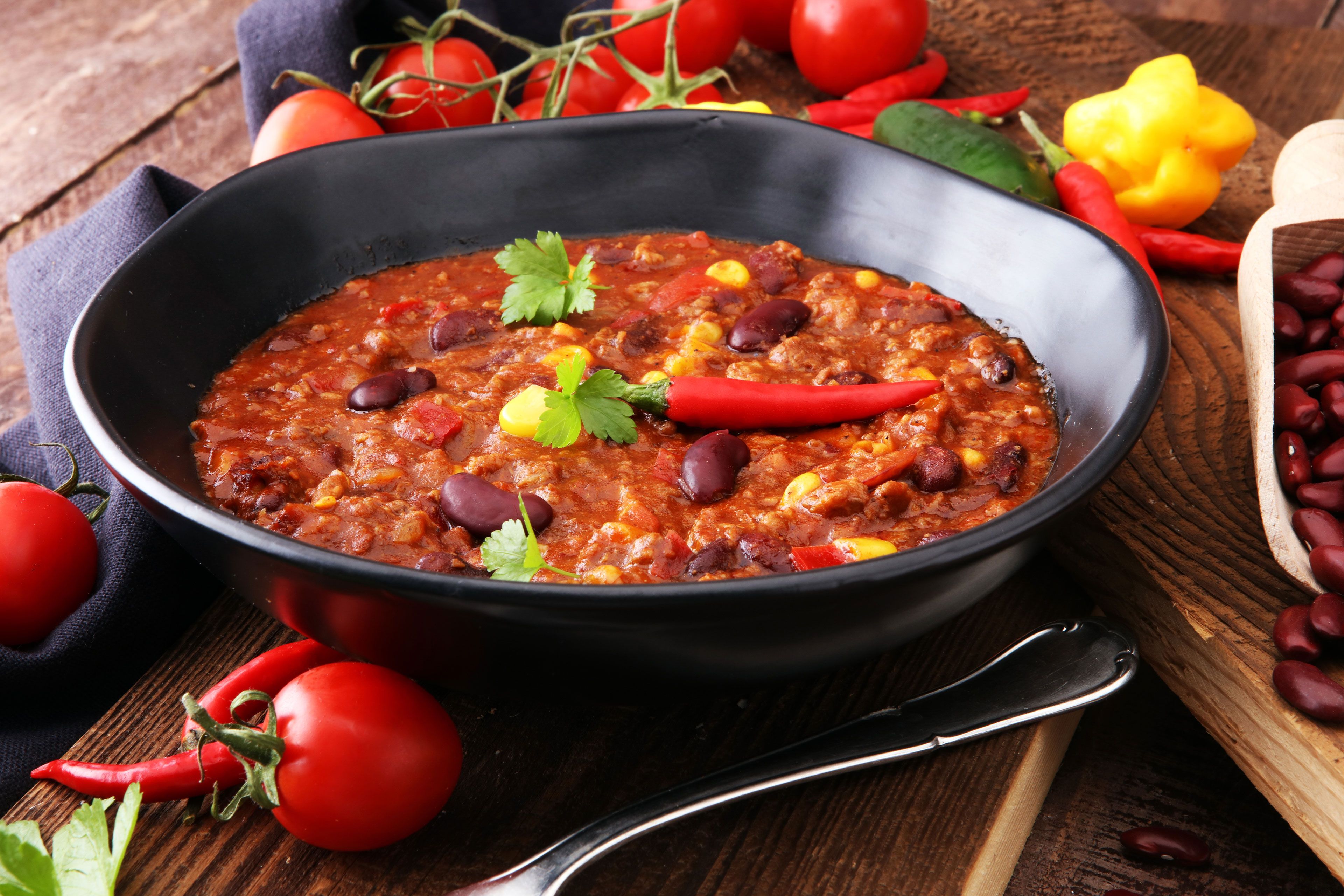 Eine Schüssel mit Chili, garniert mit roten Chilischoten und Koriander, dazu frische Tomaten und rote Kidneybohnen auf einem rustikalen Holztisch.