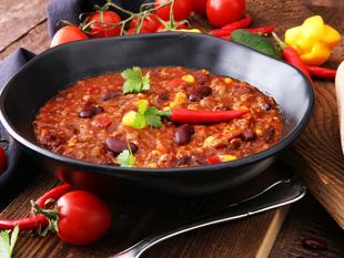 Eine Schüssel mit Chili, garniert mit roten Chilischoten und Koriander, dazu frische Tomaten und rote Kidneybohnen auf einem rustikalen Holztisch.