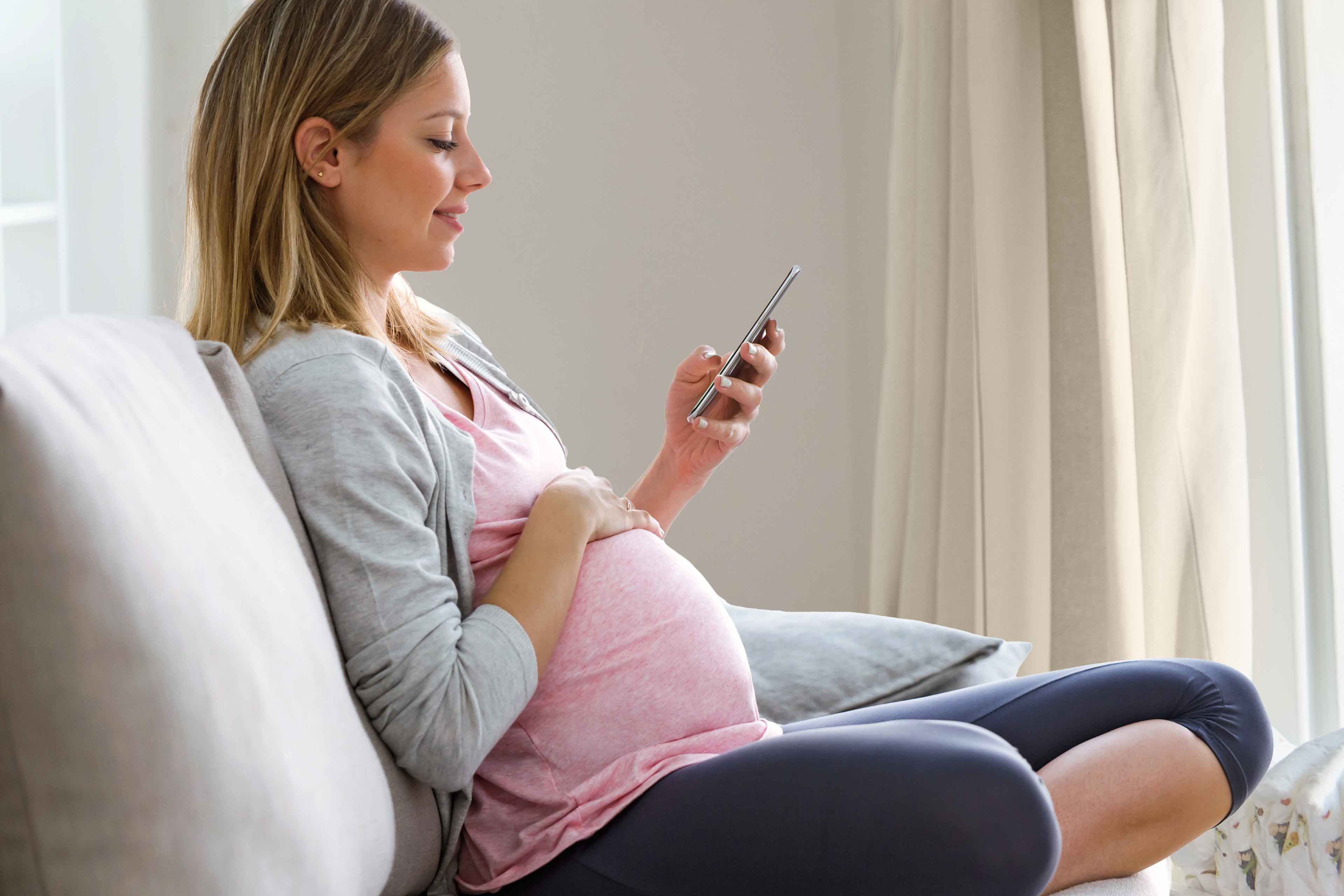 Schwangere Frau sitzt entspannt auf dem Sofa und nutzt auf ihrem Smartphone den HiPP Rechner.