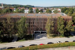 Nachhaltiges HiPP Parkhaus mit Fassade aus senkrechten Holzstämmen und begrüntem Dach, umgeben von Bäumen und Wohnhäusern.