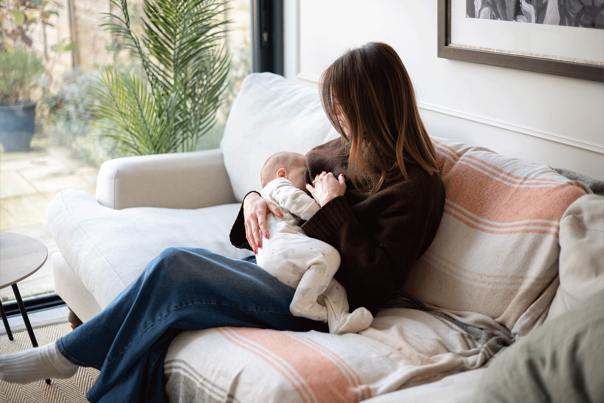 A woman with brown hair, wearing a dark brown sweater and blue jeans, sits comfortably on a white couch covered with a striped throw blanket. She holds her newborn baby, who is dressed in a white patterned onesie, gently nursing them. Next to the sofa, a large glass window reveals outdoor greenery and potted plants.