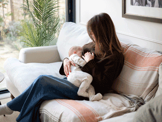 A woman with brown hair, wearing a dark brown sweater and blue jeans, sits comfortably on a white couch covered with a striped throw blanket. She holds her newborn baby, who is dressed in a white patterned onesie, gently nursing them. Next to the sofa, a large glass window reveals outdoor greenery and potted plants.