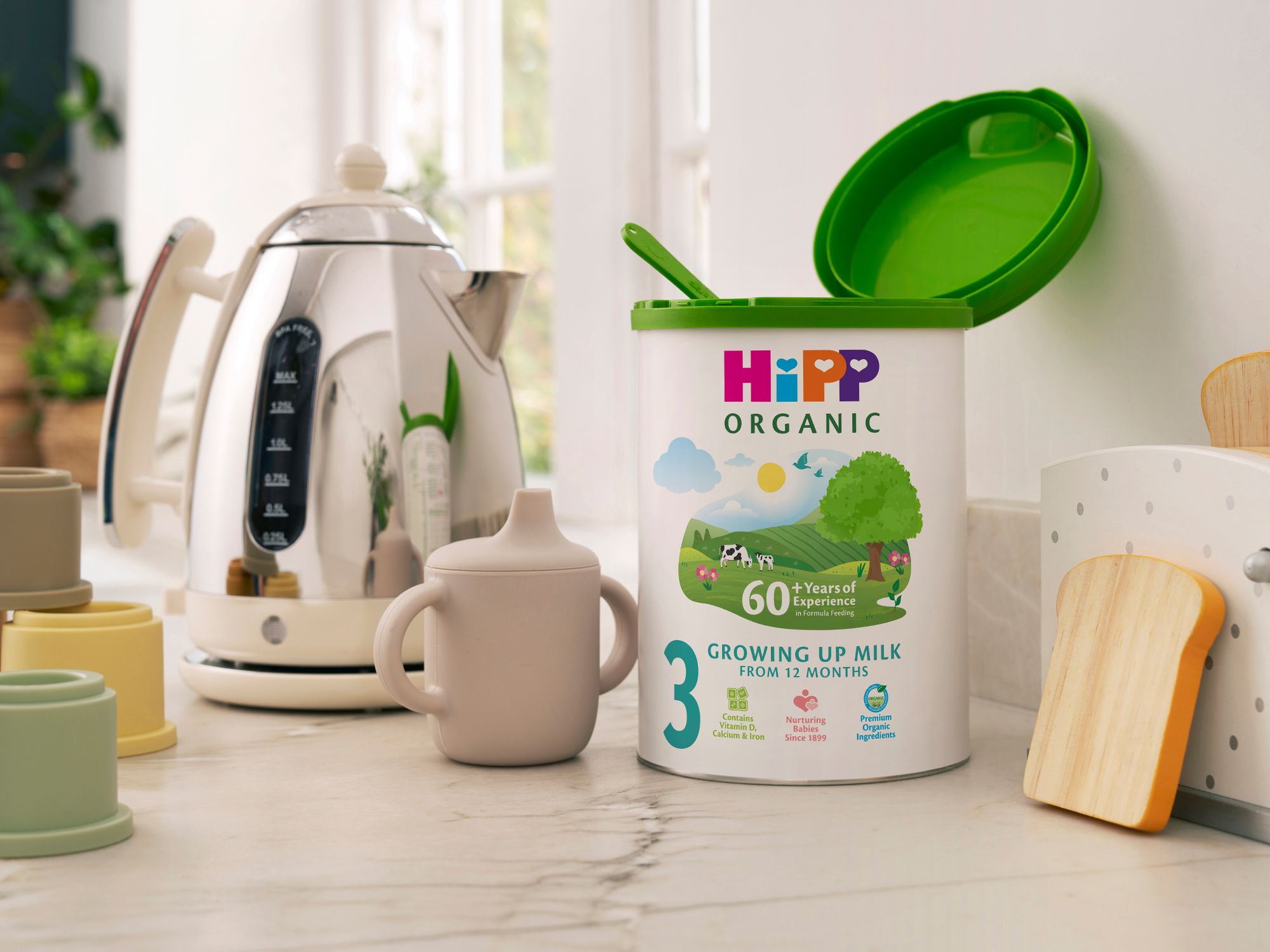 HiPP Organic Growing Up Milk Powder tin on a bright kitchen counter beside a toddler beaker and kettle, ready for milk preparation