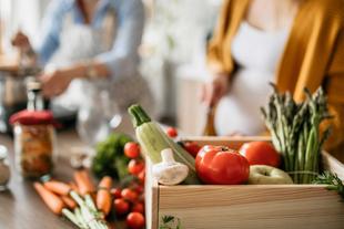 Eine Holzkiste mit frischem Gemüse, darunter Tomaten, Spargel und Knoblauch, auf einem Küchentisch mit verschiedenen Gewürzen und Utensilien im Hintergrund.