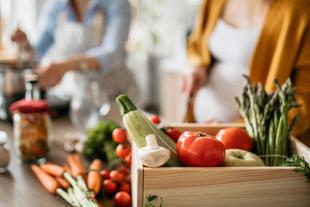 Eine Holzkiste mit frischem Gemüse, darunter Tomaten, Spargel und Knoblauch, auf einem Küchentisch mit verschiedenen Gewürzen und Utensilien im Hintergrund.