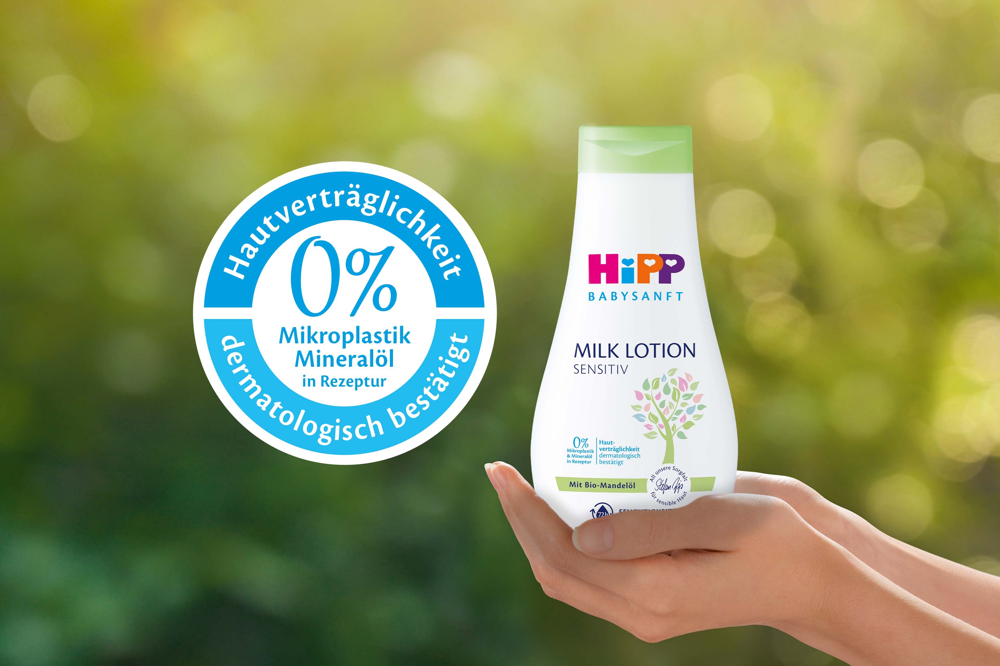 Eine Flasche eines Babypflegeprodukts mit der Aufschrift "Hipp Baby Sensitive" und einem Etikett mit der Aufschrift "0% Mikroplastik Mineralol" sowie eine Hand, die das Produkt vor einem grünen Hintergrund hält.