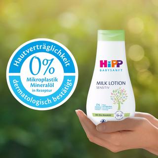 Eine Flasche eines Babypflegeprodukts mit der Aufschrift "Hipp Baby Sensitive" und einem Etikett mit der Aufschrift "0% Mikroplastik Mineralol" sowie eine Hand, die das Produkt vor einem grünen Hintergrund hält.