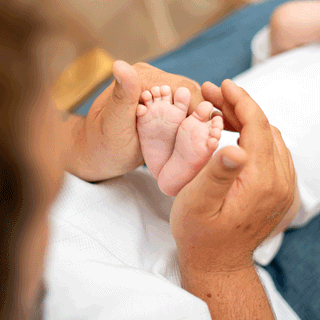 A close-up view over the shoulder of an adult softly cupping a infant's tiny feet together in the palms of their hands. The baby, dressed in a white outfit, lies in the background with arms raised near their face, slightly out of focus. The focus remains sharp on the baby's small toes and soles held securely by the parent's hands.