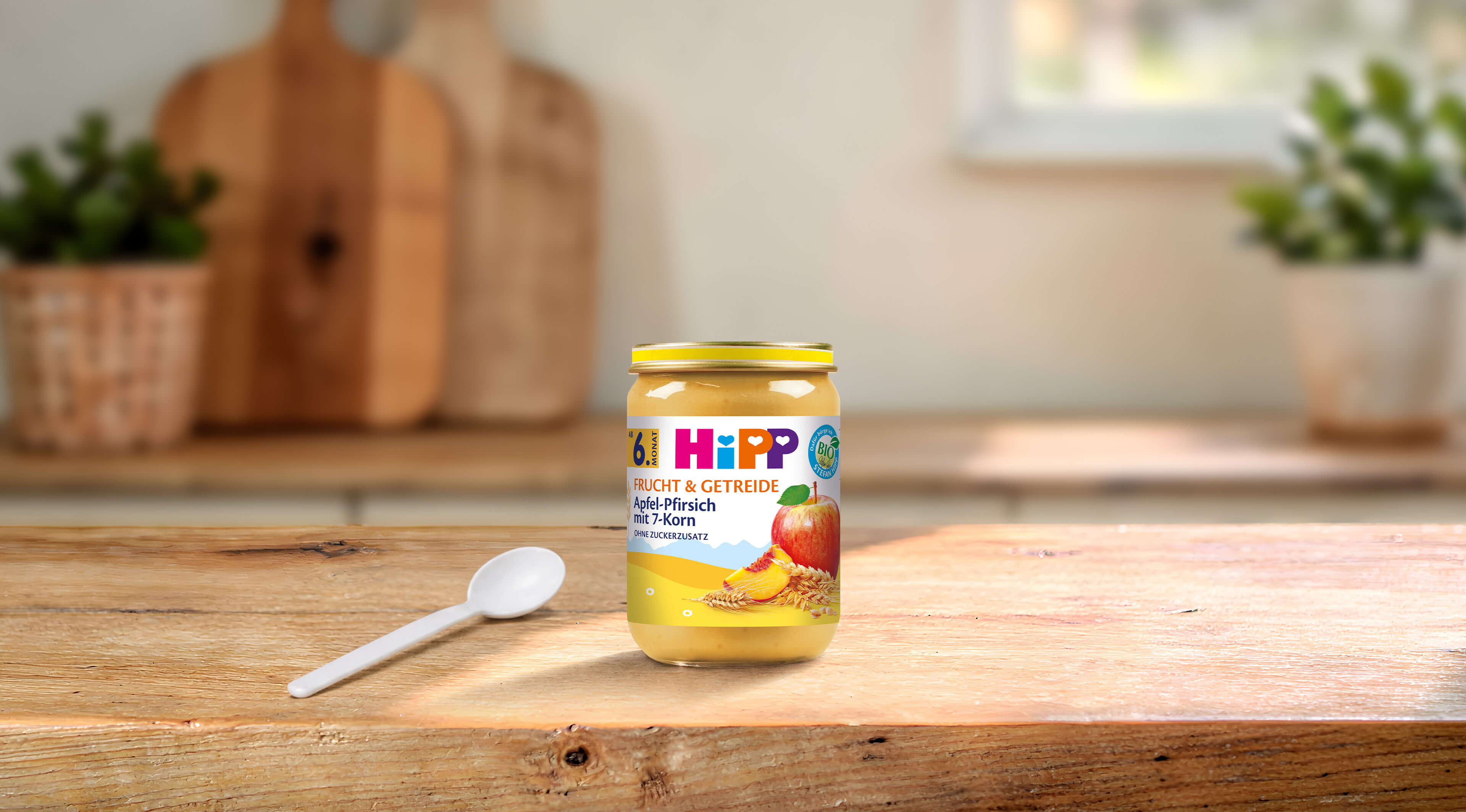 HiPP Bio-Gläschen Apfel-Pfirsich mit 7-Korn ab dem 6. Monat auf Holztisch mit Löffel in heller Küche