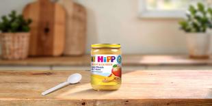 HiPP Bio-Gläschen Apfel-Pfirsich mit 7-Korn ab dem 6. Monat auf Holztisch mit Löffel in heller Küche