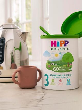 HiPP Organic Growing Up Milk tub on a bright kitchen worktop beside a toddler cup, kettle and toys, suggesting everyday family use