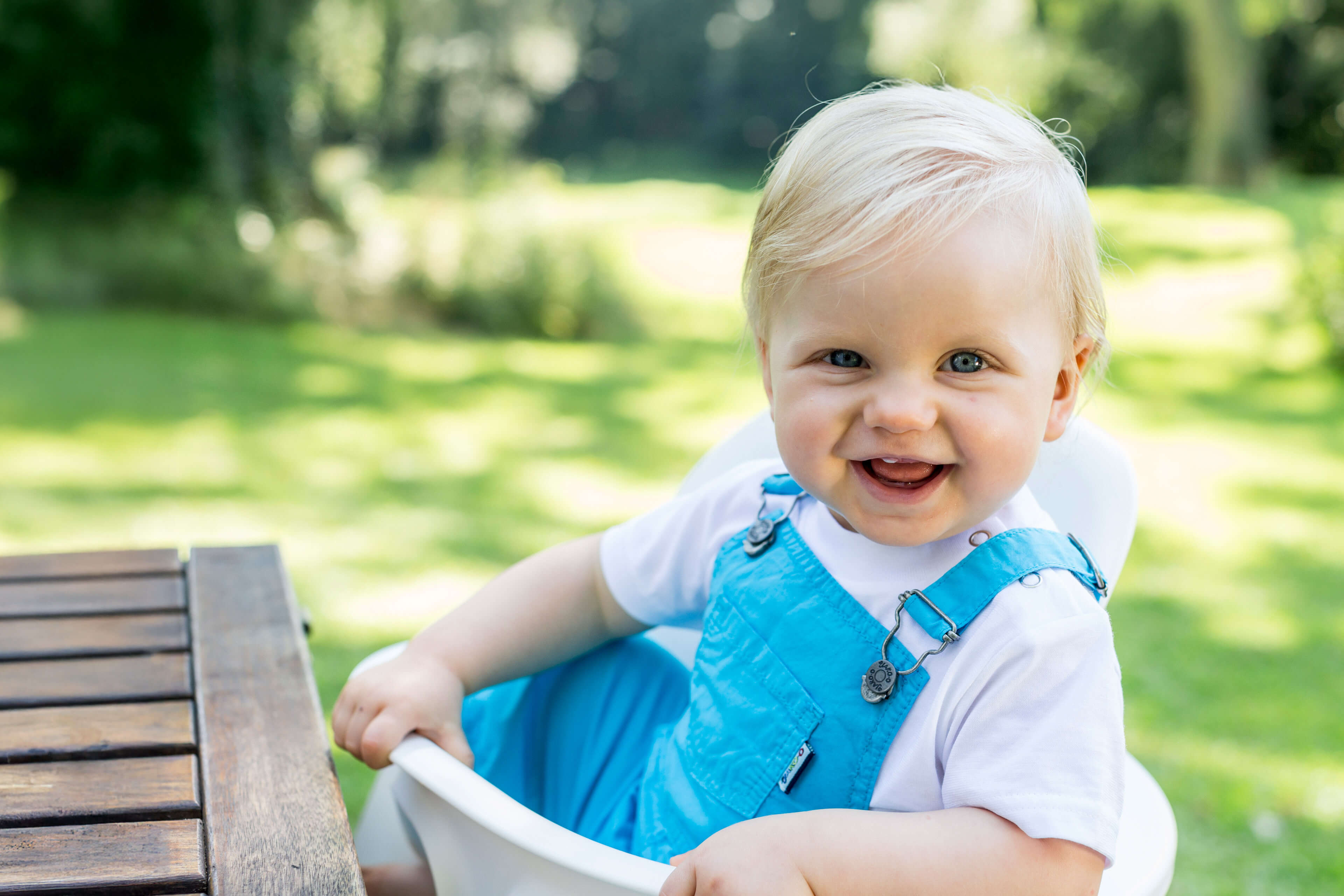 Ein fröhliches Baby in einem blauen Outfit, das im Freien vor einem unscharfen grünen Hintergrund sitzt.