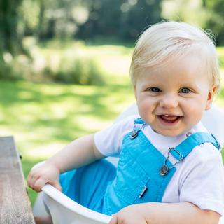 Ein fröhliches Baby in einem blauen Outfit, das im Freien vor einem unscharfen grünen Hintergrund sitzt.