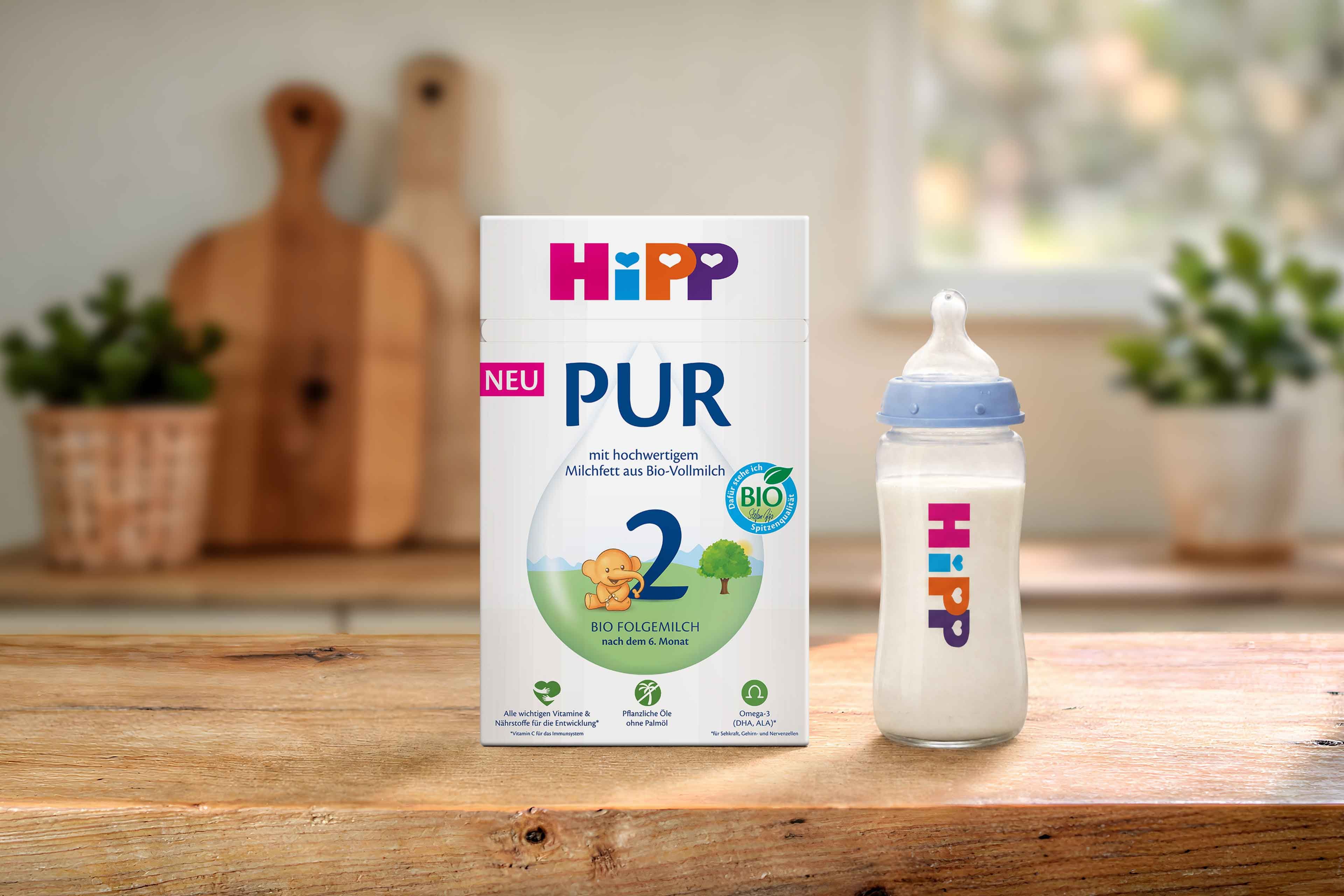 HiPP 2 PUR Bio-Folgemilch Packung mit Babyflasche auf Holztisch in heller Küche, geeignet nach dem 6. Monat