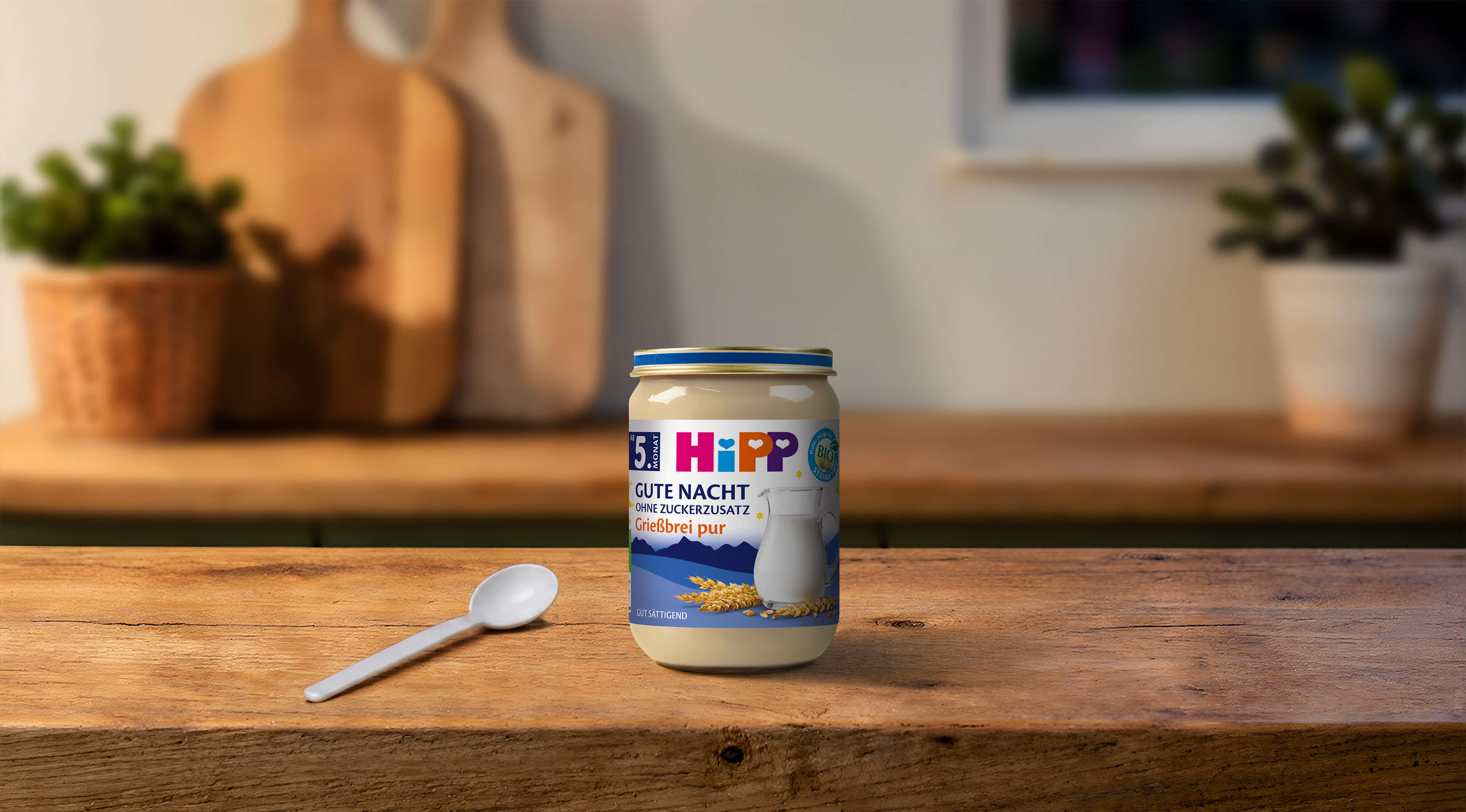 HiPP Gute Nacht Grießbrei pur im Glas auf einem Holztisch, mit Babylöffel davor in einer warm beleuchteten Küche