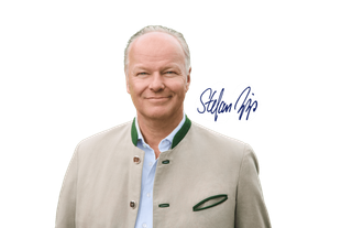 Lächelnder Mann in traditioneller, hellen Trachtenjacke vor hellem Hintergrund, daneben seine handschriftliche Unterschrift.
