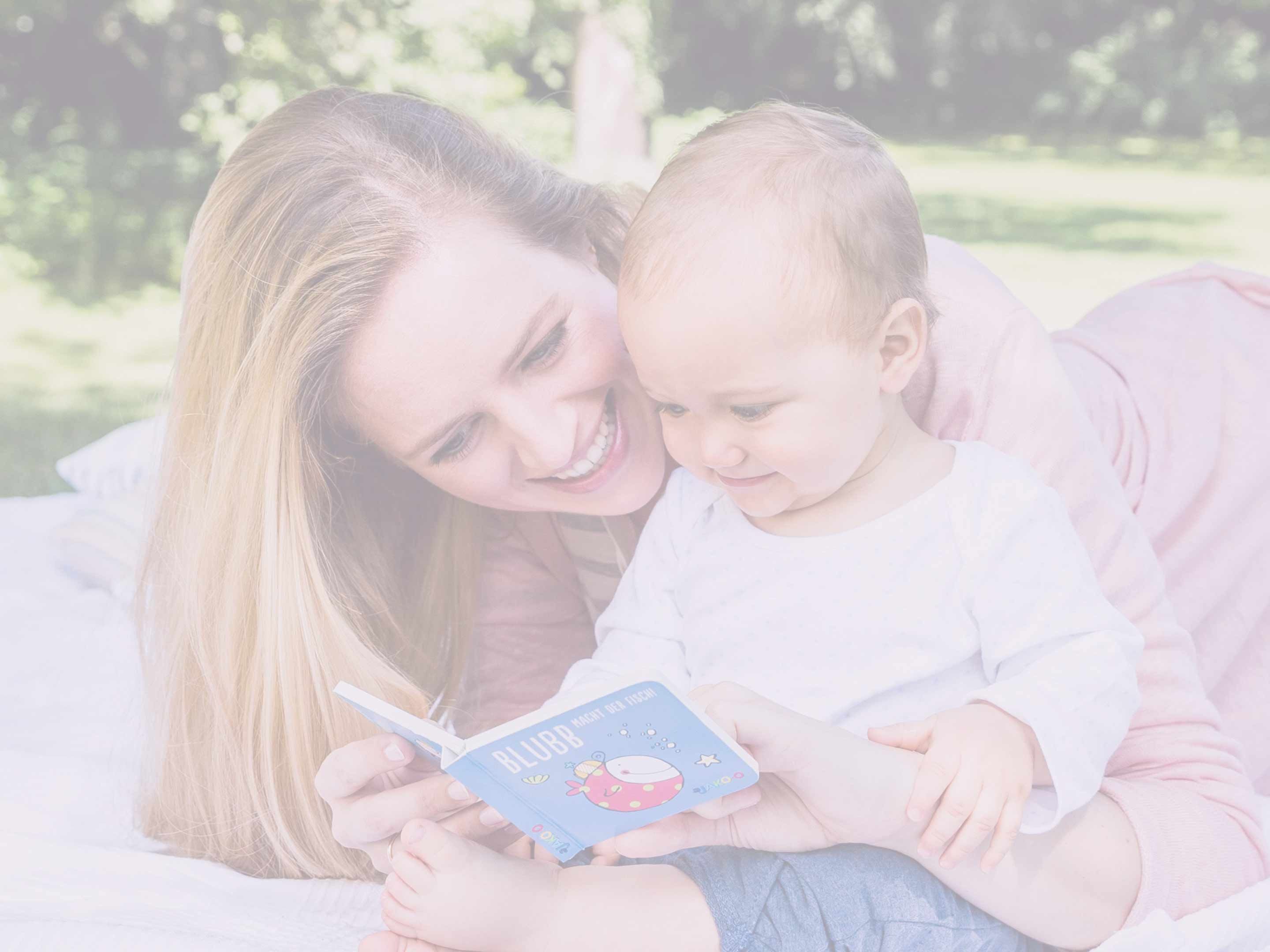 Mutter und Baby liegen im Freien auf einer Wiese, das Baby hält ein kleines blaues Buch mit einer Illustration und Text
