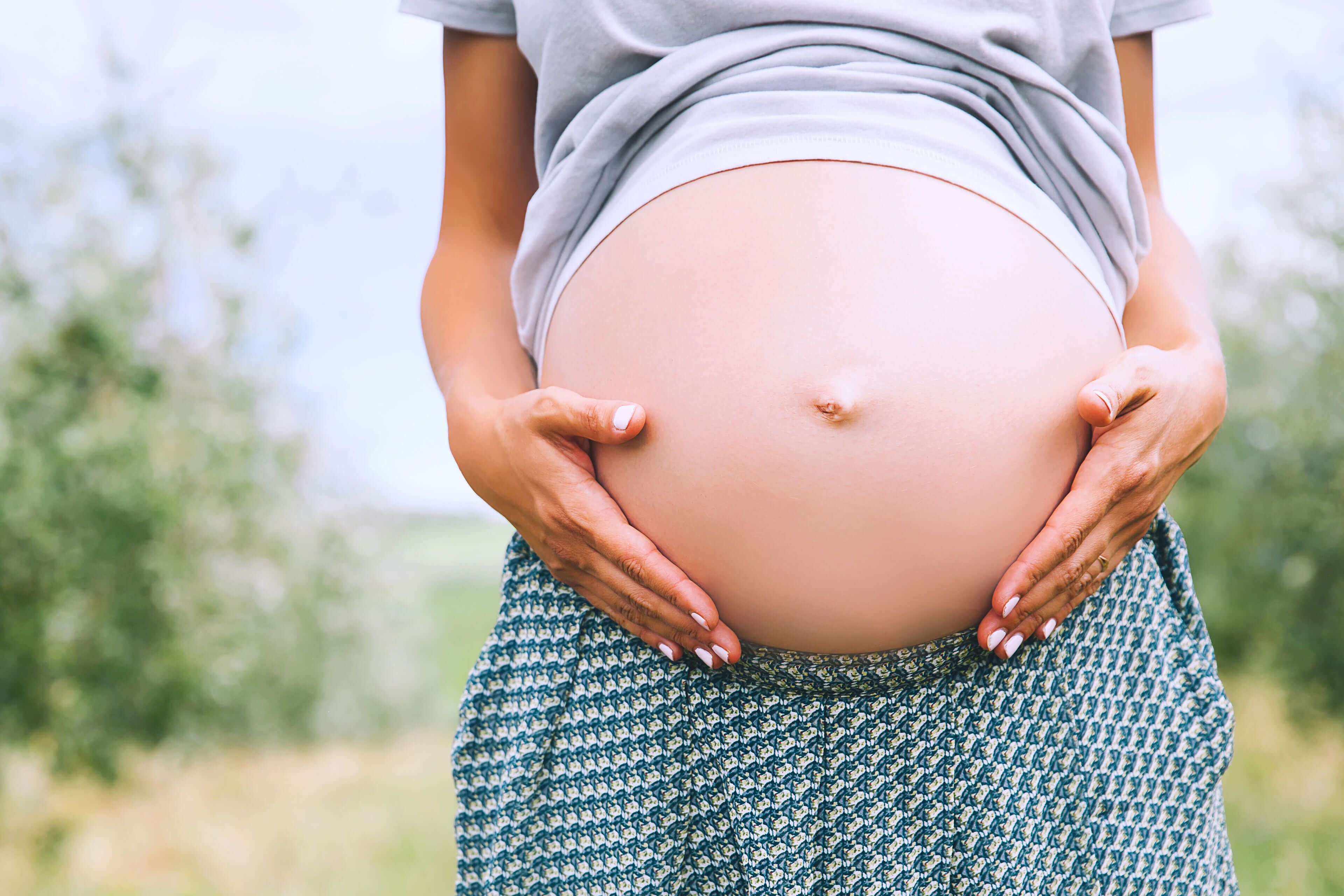 Die Hände einer Person, die sanft ihren schwangeren Bauch vor einer grünen Kulisse streichelt.