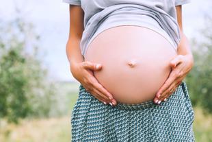 Die Hände einer Person, die sanft ihren schwangeren Bauch vor einer grünen Kulisse streichelt.