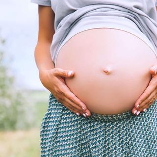 Die Hände einer Person, die sanft ihren schwangeren Bauch vor einer grünen Kulisse streichelt.