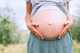 Die Hände einer Person, die sanft ihren schwangeren Bauch vor einer grünen Kulisse streichelt.
